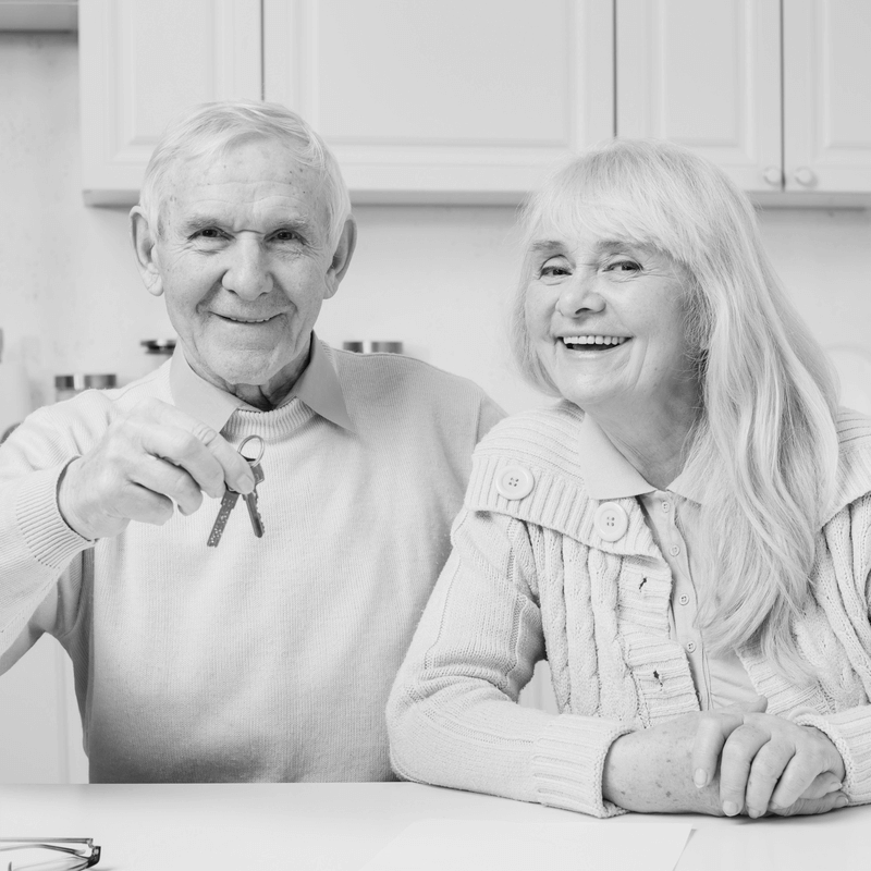 Mosolygó idős házaspár az ingatlan ajándékozási szerződéskötés után, kezükben a lakás kulcsaival. Az idős férfi büszkén mutatja a kulcsokat, míg felesége boldogan mosolyog mellette. A konyhában ülnek, meghitt és bizalommal teli légkörben, amely az ingatlan ajándékozási szerződés nyugodt lebonyolítását sugallja. A kép szimbolizálja az ingatlan vagy egyéb vagyon átadását szeretett személyeknek.