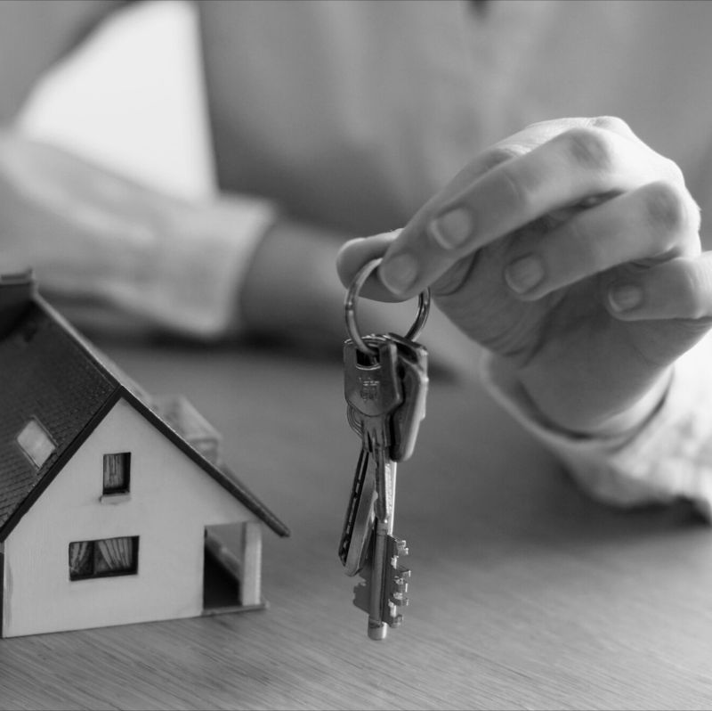 Egy kéz kulcscsomót tart egy asztalon lévő házmodell felett, amely a haszonélvezeti jog bejegyzését vagy törlését szimbolizálja. A kép az ingatlan tulajdonjogának és használati jogának átadását jelképezi, utalva a jogi folyamatokra, például a haszonélvezeti jog bejegyzésére vagy törlésére. Az elmosódott háttér hivatalos ügyintézést sugall.