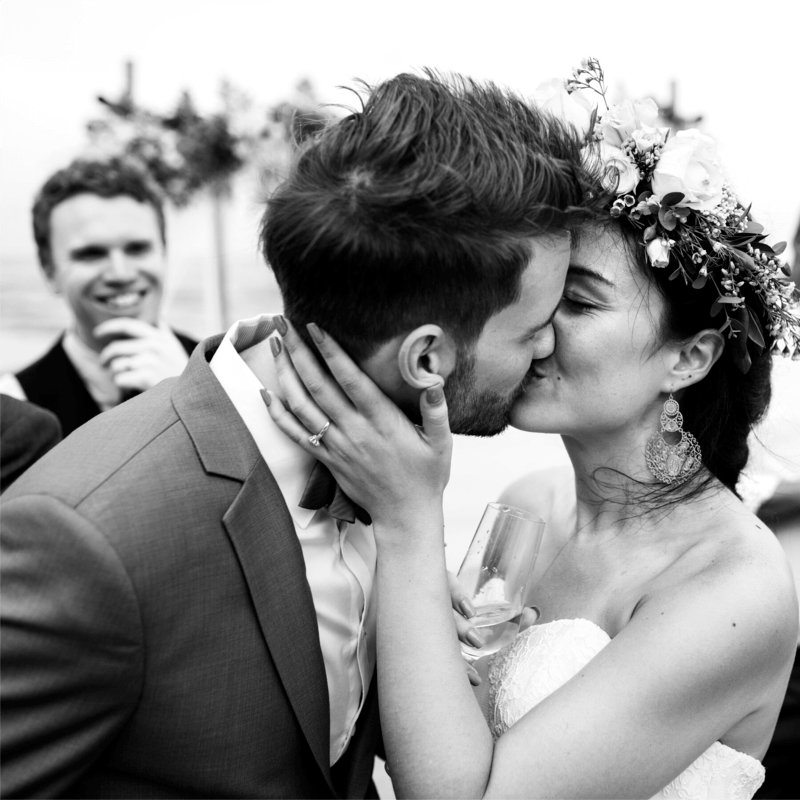 Friss házaspár boldog csókja az esküvő után, miközben a menyasszony kezében pezsgőspoharat tart. A nő virágkoronát visel, és eljegyzési gyűrűje látható a kezén, ahogy gyengéden megérinti férje arcát. A háttérben mosolygó vendégek figyelik a pillanatot, ünnepi hangulatot teremtve. A kép a házassági és élettársi vagyonjogi szerződéskötés fontosságát is szimbolizálhatja, amely a közös élet és a vagyoni viszonyok biztonságát teremti meg.