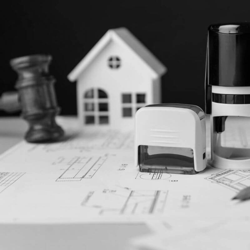 Ingatlanjogi dokumentumok és pecsétek egy asztalon, a közös tulajdon megszüntetésével kapcsolatos ügyintézést jelképezve. A tervrajzokon hivatalos bélyegzők, egy kis házmodell és egy bírói kalapács látható, amelyek az ingatlan tulajdonjogának változását szimbolizálják. A kép a közös tulajdon megosztását, eladását vagy jogi rendezését illusztrálja.