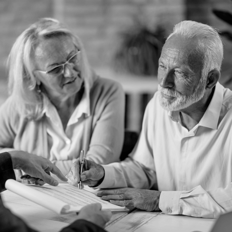 Idős házaspár végrendelet és öröklési szerződéskötés során, ügyvéd jelenlétében. Az elegánsan öltözött idős férfi aláír egy hivatalos dokumentumot, miközben felesége figyelmesen követi a folyamatot. Az ügyvéd mutatja a megfelelő helyet az aláírásra, segítve az öröklési szerződés lebonyolítását. Barátságos, professzionális környezet, amely a jogi ügyintézés bizalmi jellegét hangsúlyozza.