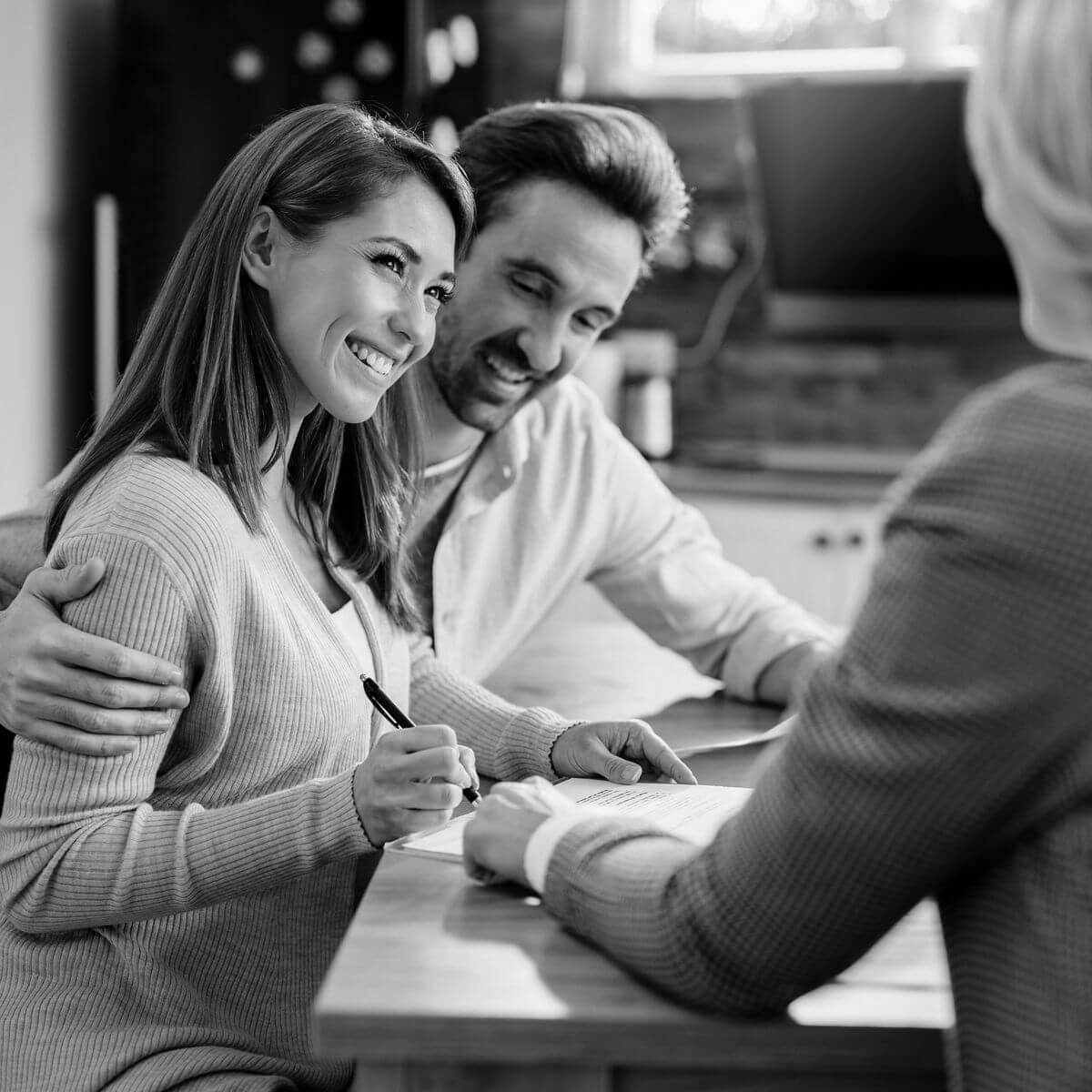 Fiatal pár mosolyogva írja alá az ingatlan adásvételi szerződést egy ügyvéd jelenlétében – bizalommal és jogi biztonsággal zajló lakásvásárlás.