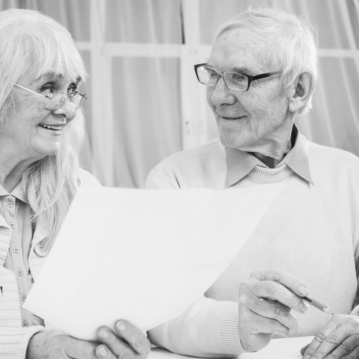 Idősebb házaspár ingatlan ajándékozás dokumentumait nézi át Idős nő és férfi együtt néznek egy dokumentumot és mosolyognak – ingatlan ajándékozás folyamatát átbeszélő, közös adminisztrációs pillanat.