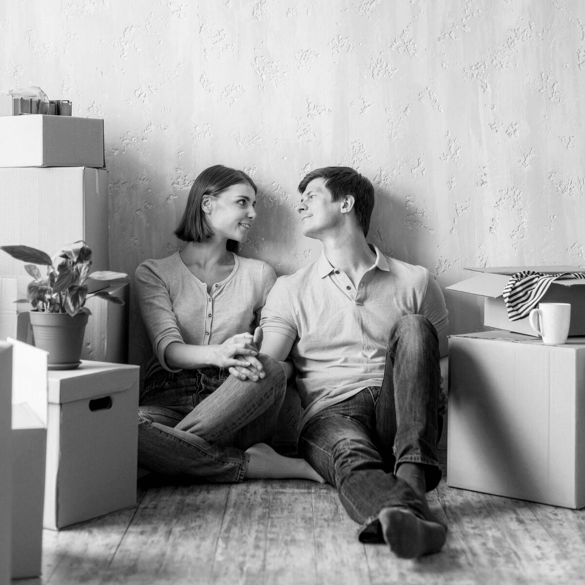 Fiatal pár ül a földön, mosolyogva egymásra néznek egy új lakásban, körülöttük kartondobozok és egy növény. A kép jól illusztrálja az otthon start hitel igénylésével kapcsolatos lakásvásárlás örömét és a költözés izgalmát.