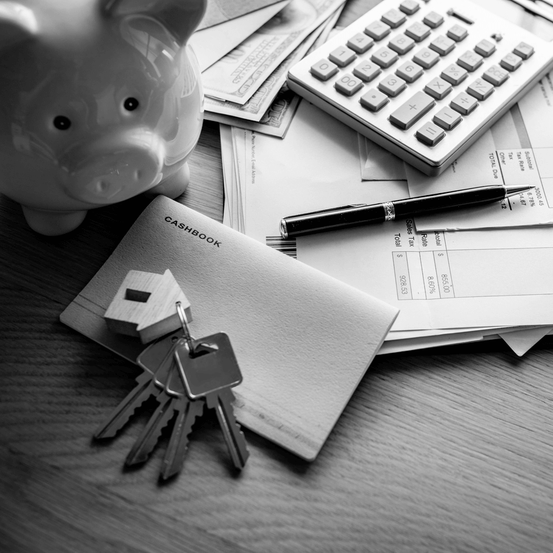 Ingatlan adásvétel illetéke és költségei, illusztrálva ház alakú kulcstartóval és kulcsokkal, pénzügyi dokumentumokkal, készpénzzel, számológéppel és persellyel az asztalon.