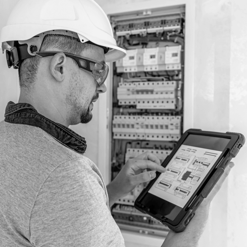 Villanyszerelő védősisakban és védőszemüvegben tablet segítségével ellenőrzi egy ingatlan elosztószekrényének elektromos kapcsolási rajzát, az ingatlan adásvétel előtti műszaki állapotfelmérés részeként.