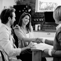 ingatlan-adasvetel-ugyvedi-segitseggel Fiatal pár mosolyogva kezet fog ügyvéddel egy konyhaasztalnál, miközben ingatlan adásvételi szerződést írnak alá – a kép az ingatlanjogi ügyvédi szolgáltatások biztonságot nyújtó szerepét ábrázolja az otthonvásárlás során.