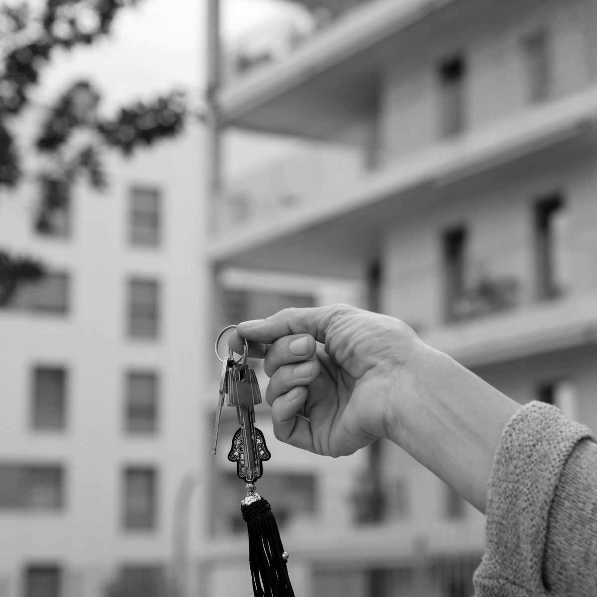 Közeli kép egy kézről, amely egy kulcscsomót tart, mögötte egy modern társasház látható, a bérleti szerződés átadását szimbolizálva.