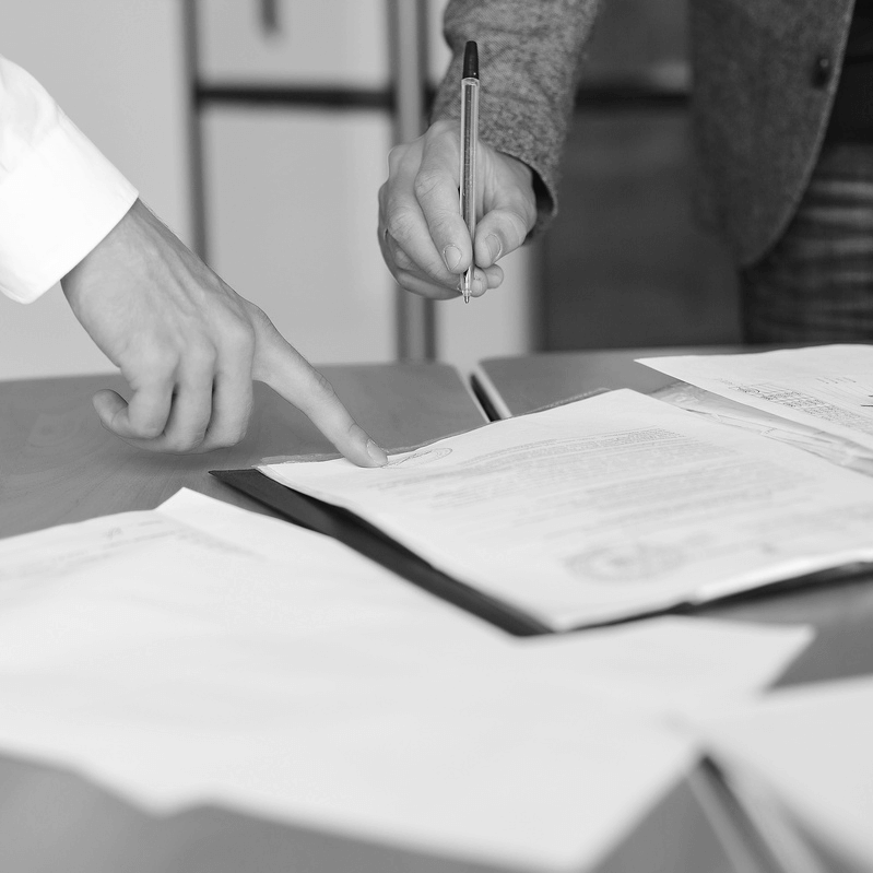 Két személy egy bérleti szerződés részleteit egyezteti és aláírásra készíti elő, az egyik kéz a dokumentum fontos pontjára mutat, míg a másik tollal a szerződés aláírására készül.