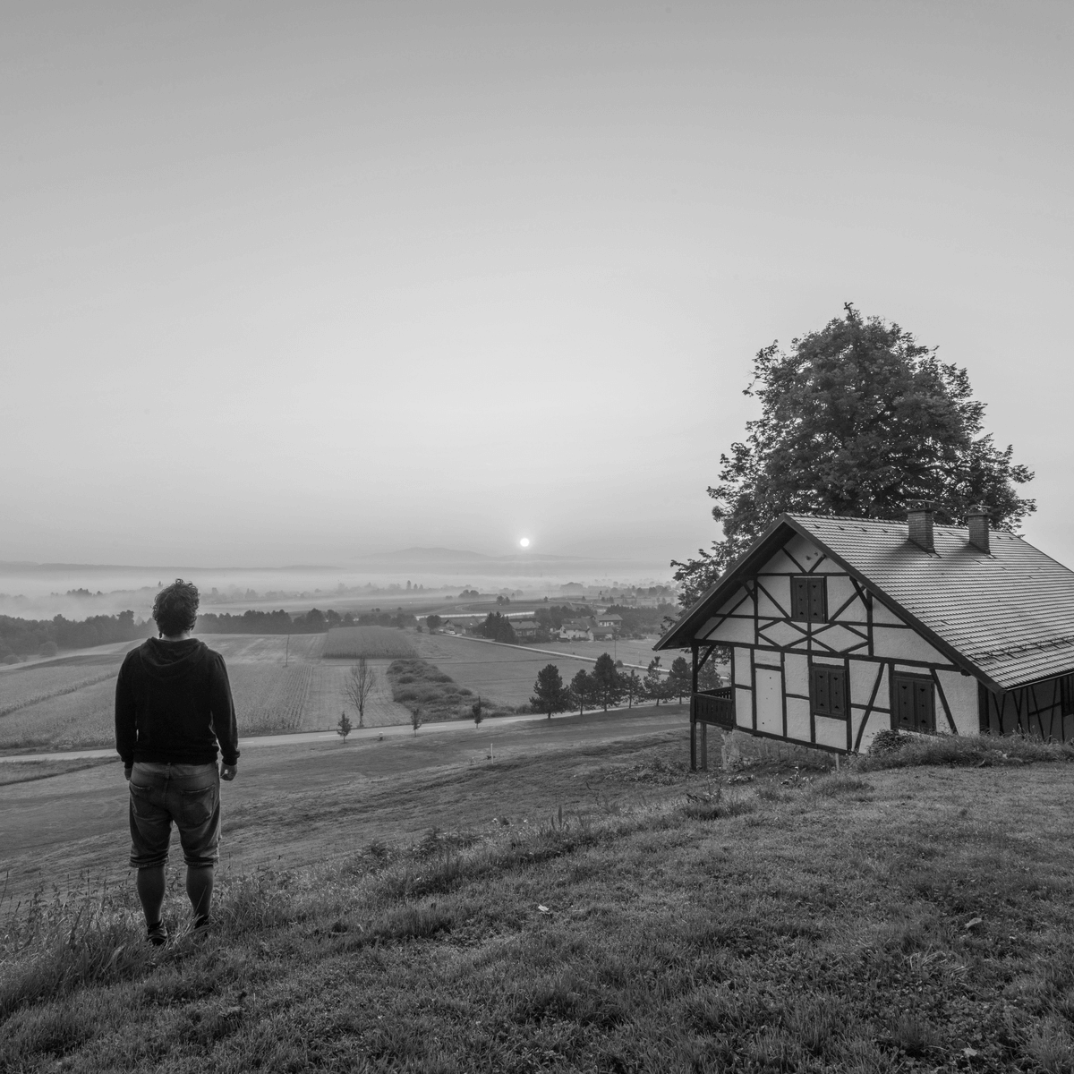 Zártkerti környezetben álló vidéki ház domboldalon, előtérben egy ember a tájat szemlélve, háttérben mezők és napfelkelte – a zártkert átminősítése utáni beépíthetőség hangulatát bemutató fotó.